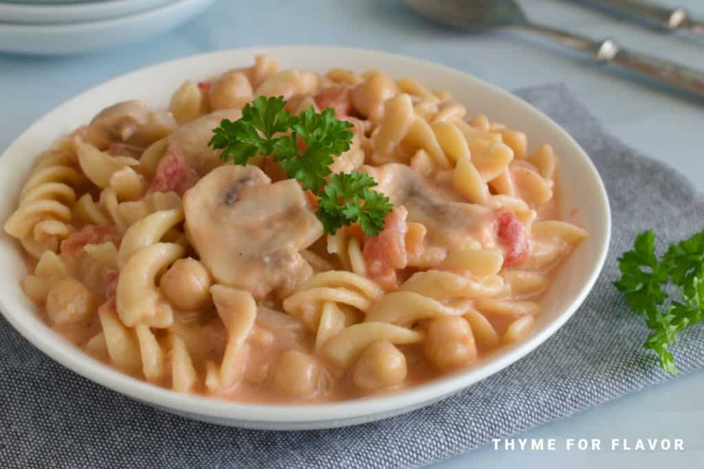A bowl of creamy pasta with chickpeas, mushrooms, and a garnish of fresh parsley. The dish is placed on a gray cloth with the text "THYME FOR FLAVOR" in the bottom right corner. A fork and spoon are partially visible in the background.