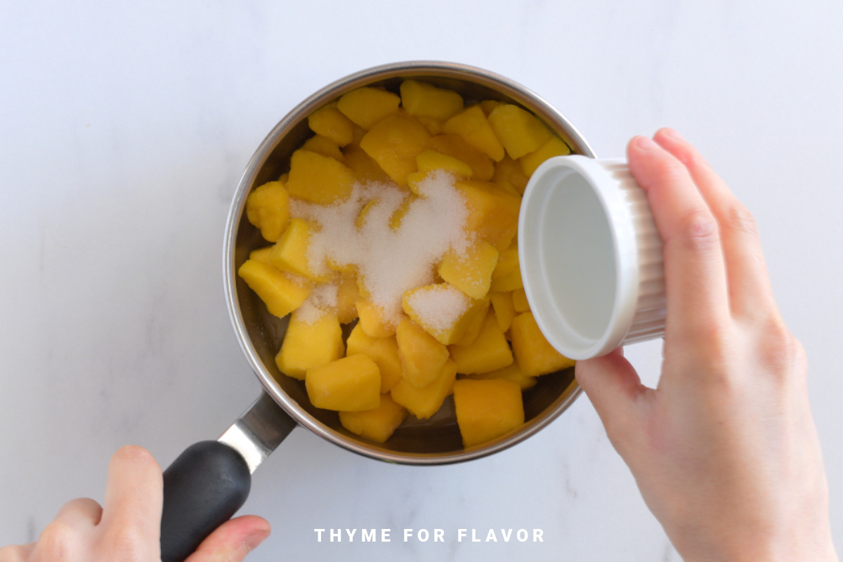Placing sugar into a saucepan with mango pieces in it.