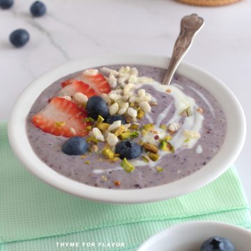 Close up image of purple smoothie bowl with a spoon, sliced strawberries, blueberries, puffed rice, and chopped pistachios.
