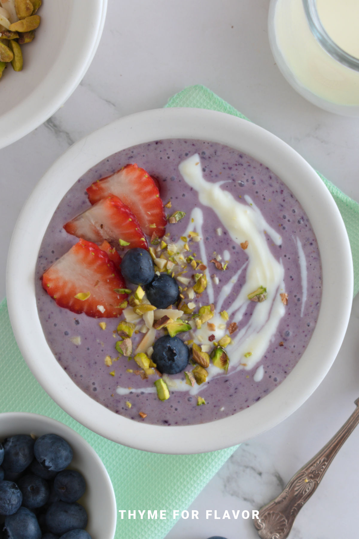 Top shot image of a purple smoothie bowl in a white bowl, topped with strawberries, blueberries, pistachios, and yogurt.