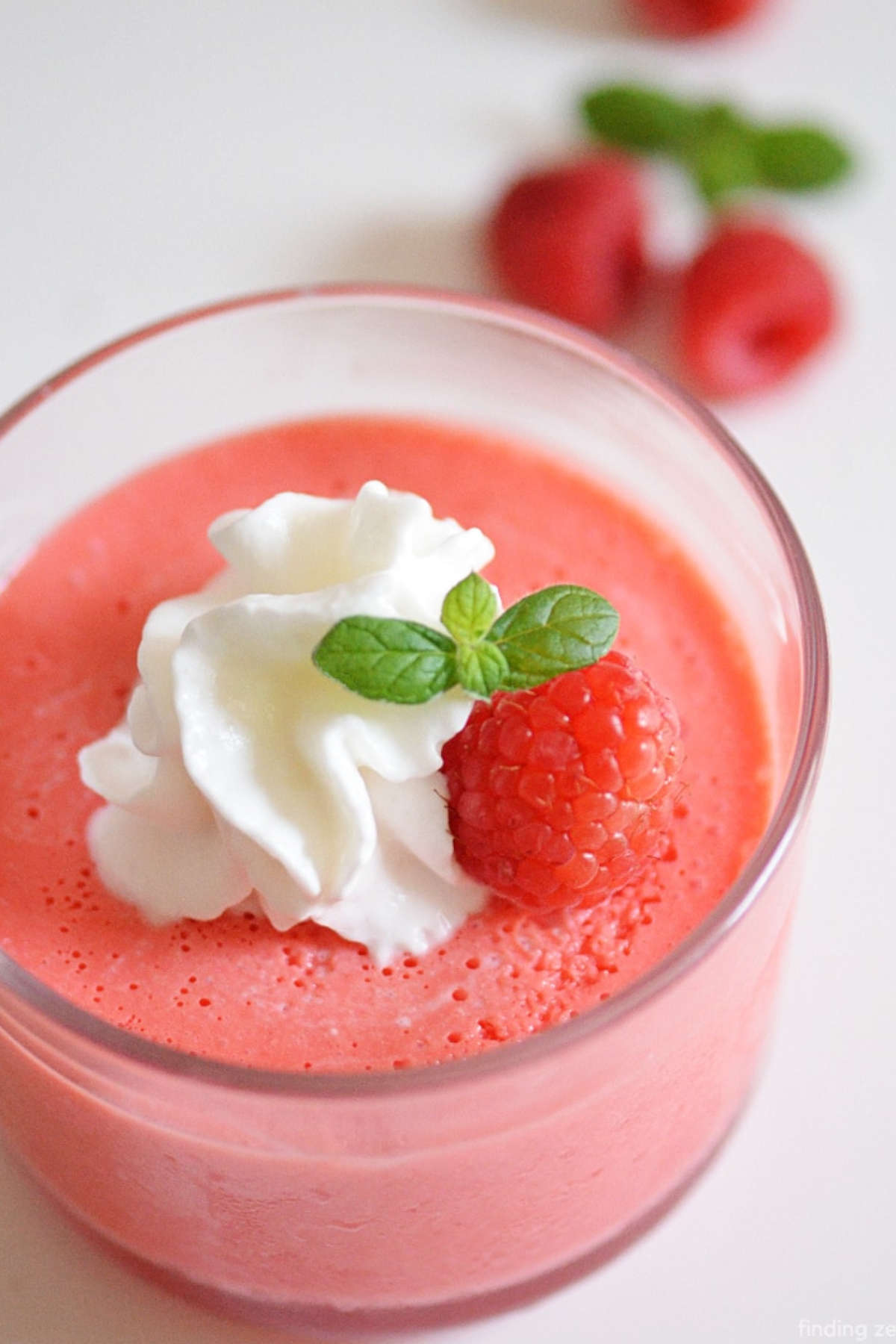 Close up image of pink jello in a glass, topped with whipped cream, mint leaves, and a raspberry.